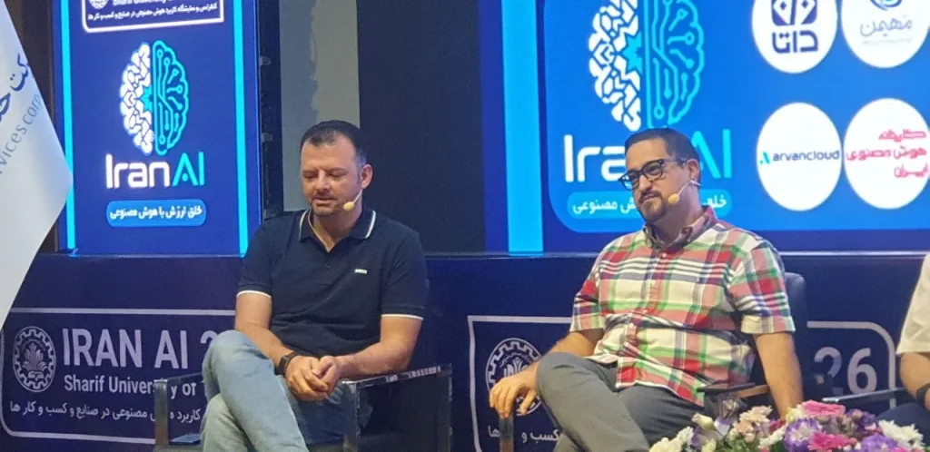Two speakers seated onstage beside Iran AI signage.