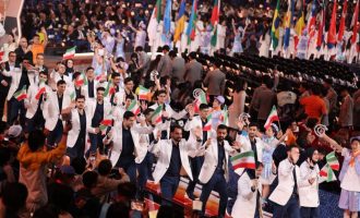 Group in light jackets waving Iranian flags along an arena walkway.