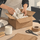 Hands packing a mug in a cardboard box beside other goods and parcels. Conceptual editorial illustration.