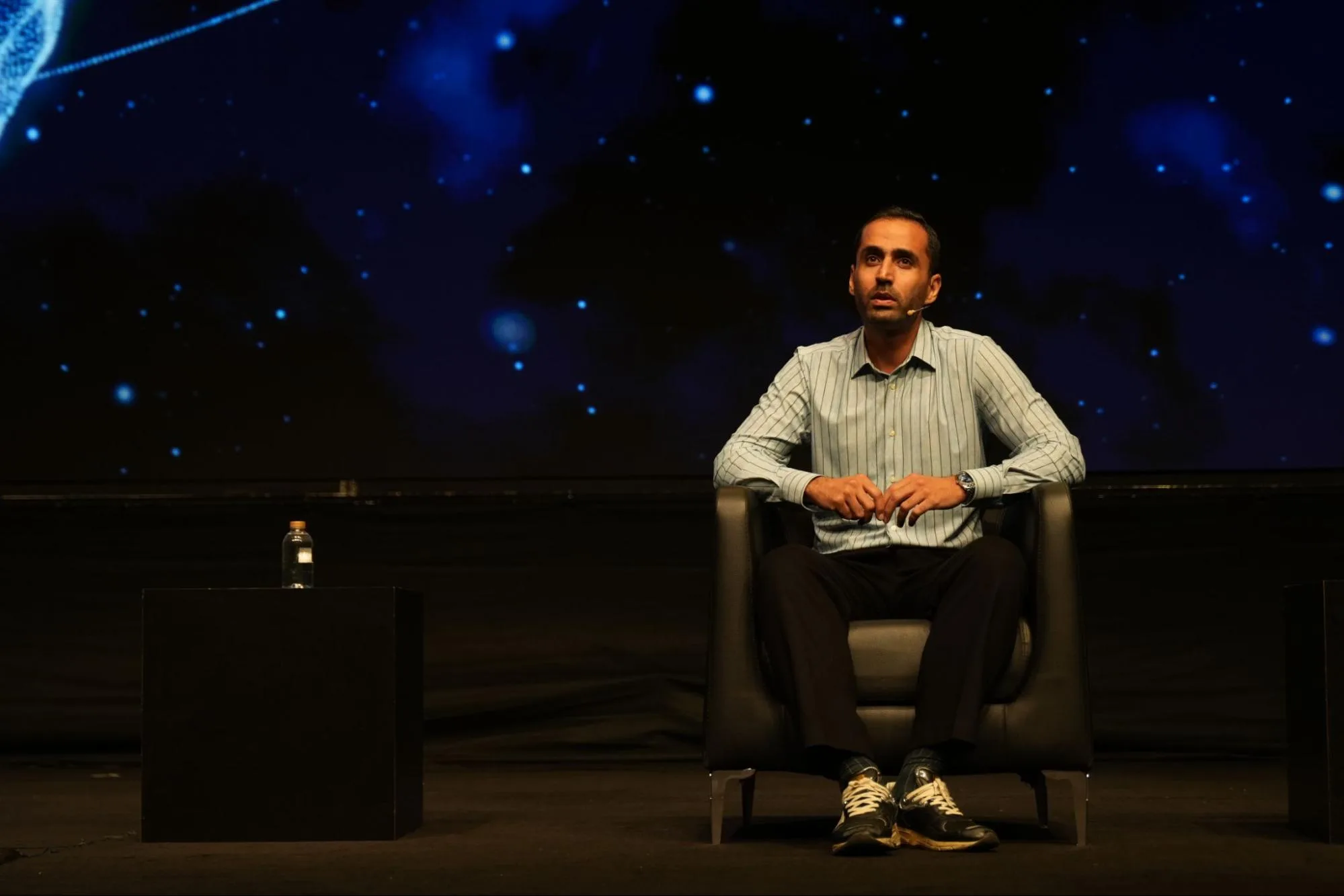 A man in a striped shirt sits onstage in front of a blue event screen.