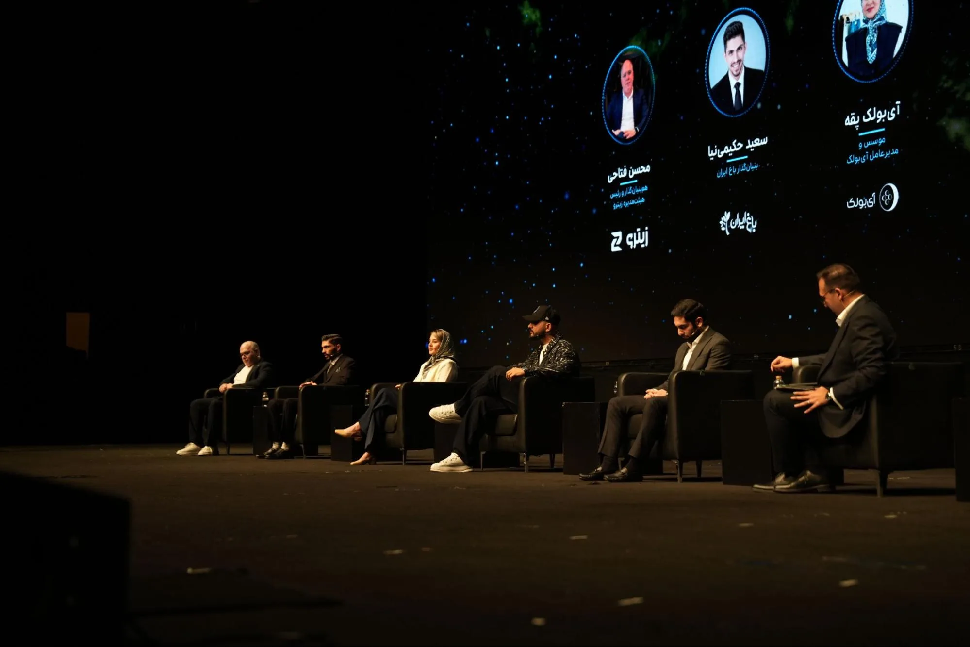 Six participants sit onstage beneath a screen displaying speaker portraits.