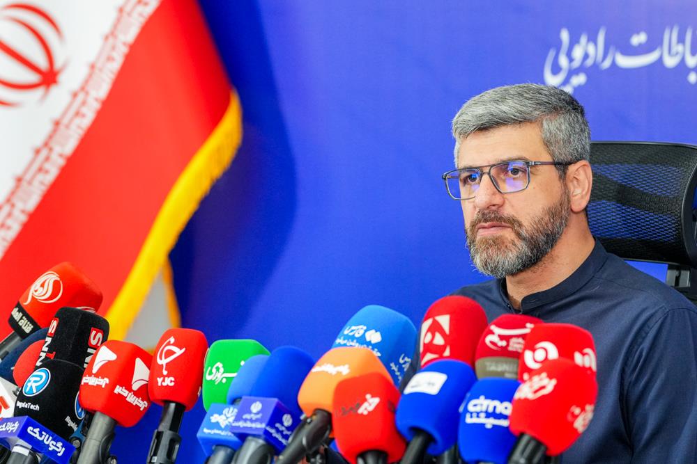 Hamid Fattahi at a press conference, with microphones in front of him and an Iranian flag beside him.