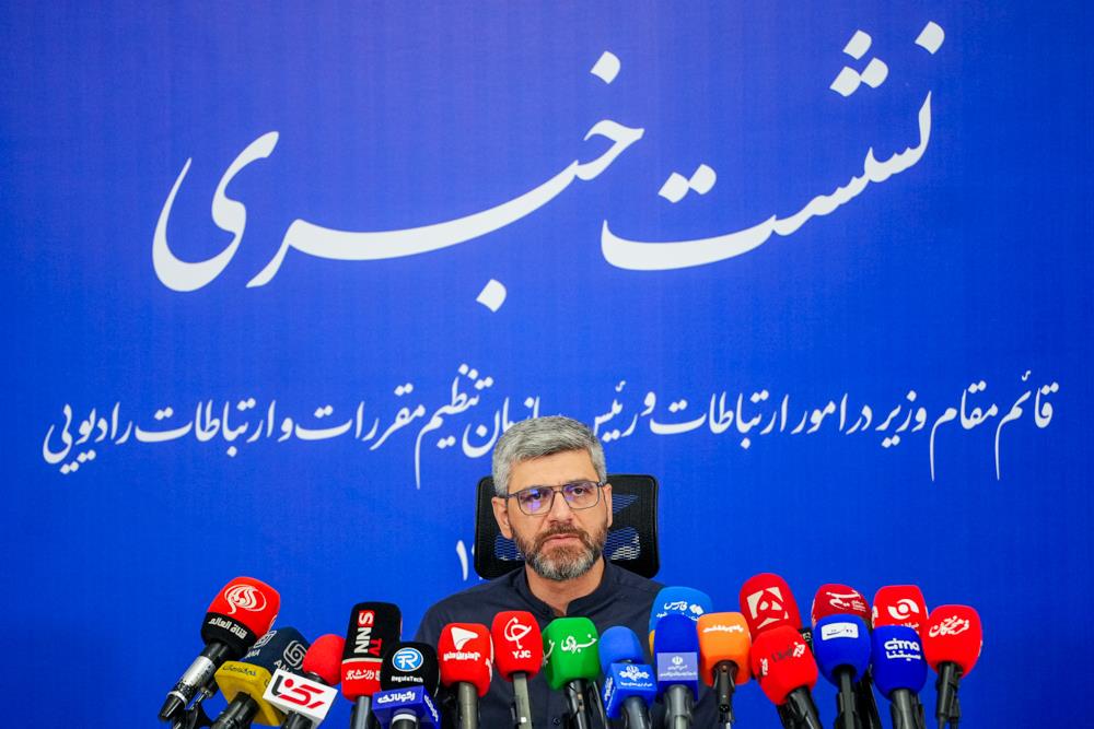 Hamid Fattahi behind a row of microphones beneath a blue press conference backdrop.