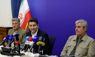 Sattar Hashemi sits between two men at a table crowded with microphones, with an Iranian flag behind them.