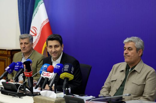 Sattar Hashemi sits between two men at a table crowded with microphones, with an Iranian flag behind them.