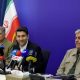 Sattar Hashemi sits between two men at a table crowded with microphones, with an Iranian flag behind them.