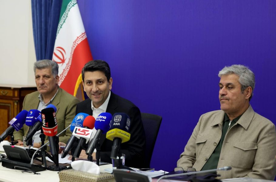 Sattar Hashemi sits between two men at a table crowded with microphones, with an Iranian flag behind them.