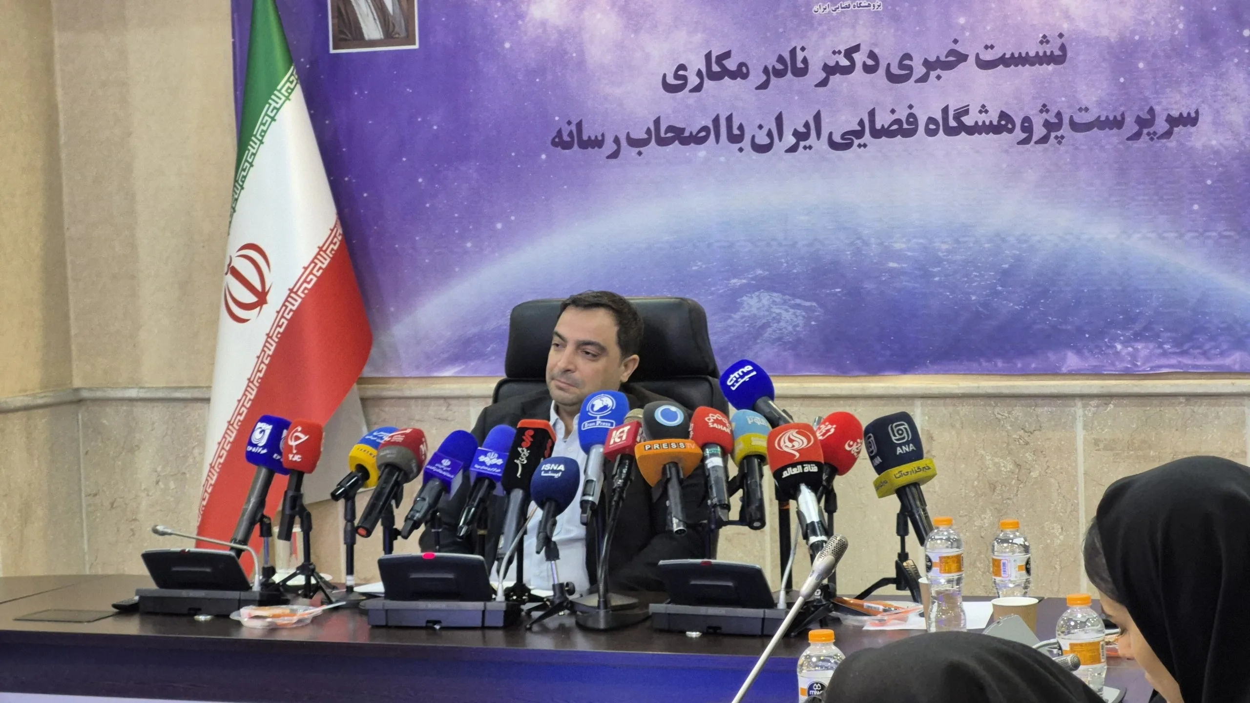A speaker sits behind a bank of microphones beneath a Persian banner naming Nader Makari and the Iranian Space Research Center.